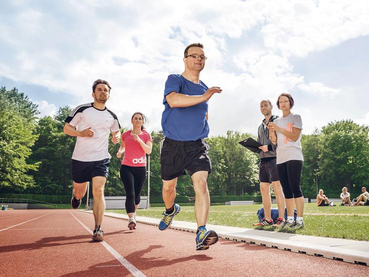 Inklusive Laufgruppe mit Läufer mit Prothese auf der Bahn; zwei Personen beobachten mit Stoppuhr und Klemmbrett.